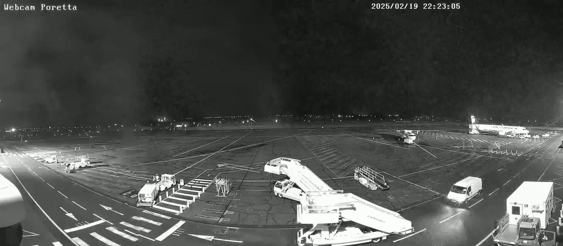 A nighttime, overcast view of an airport tarmac showing several planes, ground support vehicles, and service trucks.