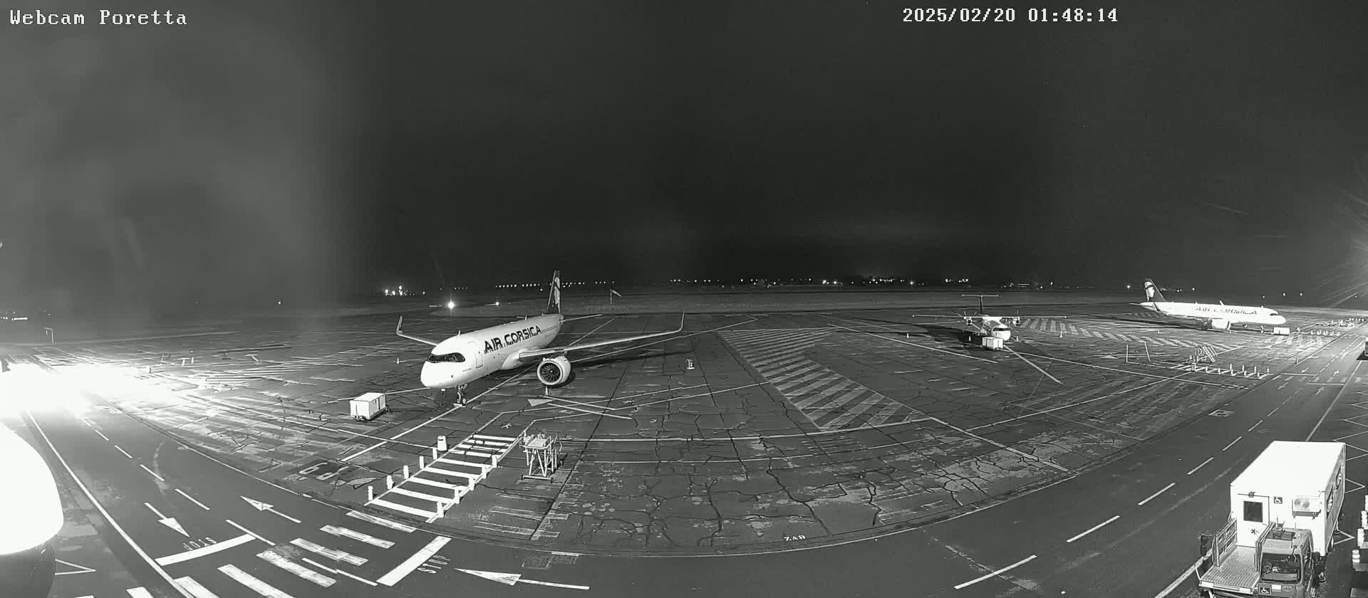 At night, on a tarmac, two airplanes are parked, one of which is an Air Corsica plane; a ground service vehicle is visible in the lower right corner.