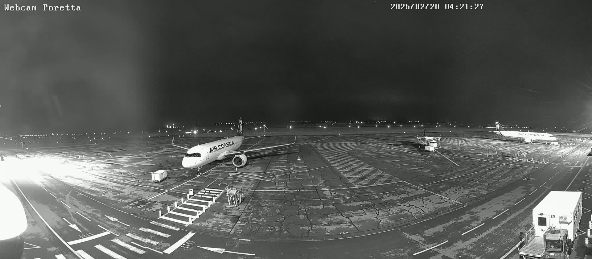 A nighttime, black and white view of an airport tarmac showing two airplanes parked and various ground support vehicles.
