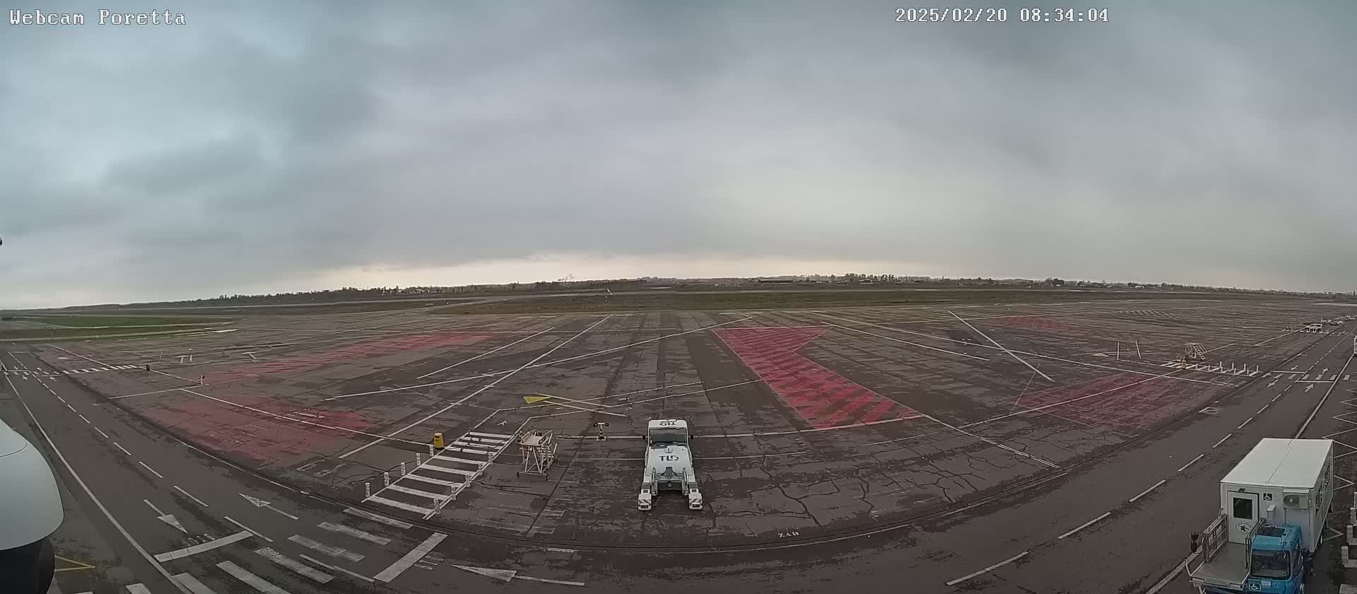 An overcast sky covers a large, empty airport tarmac with a few service vehicles and markings on the ground.