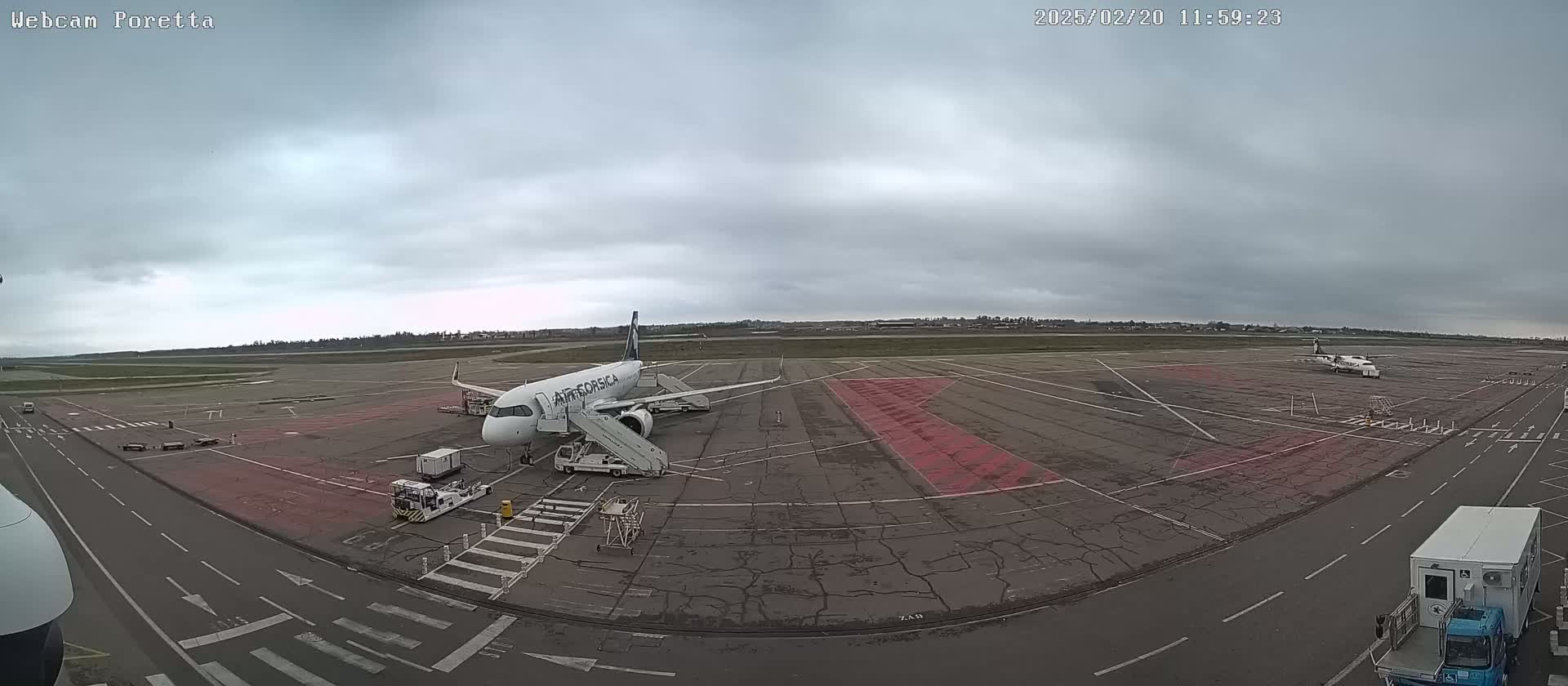 An airplane is parked at an airport tarmac on an overcast day, with ground support equipment nearby and another smaller plane visible in the distance.