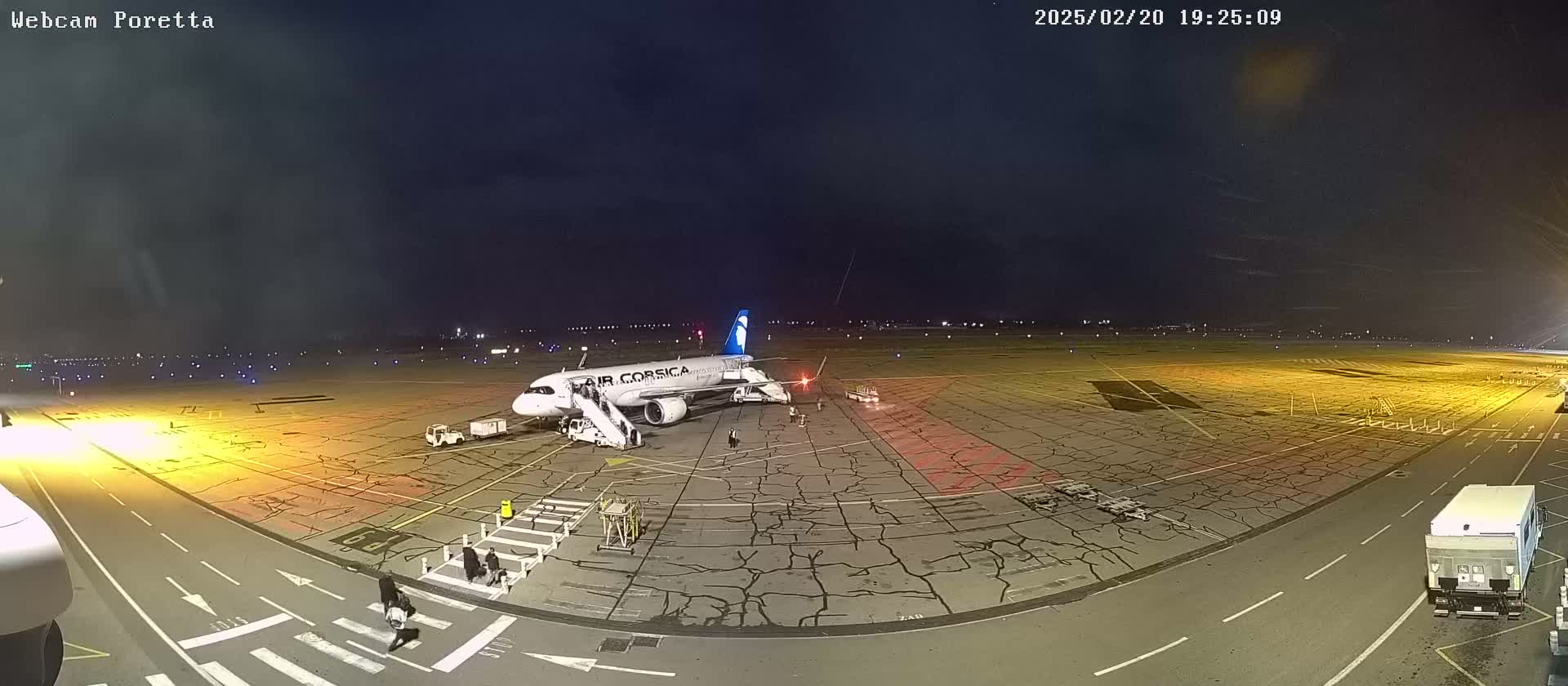 At night, an airplane is parked on a tarmac with several people and ground support vehicles nearby, under a dark, possibly cloudy sky.