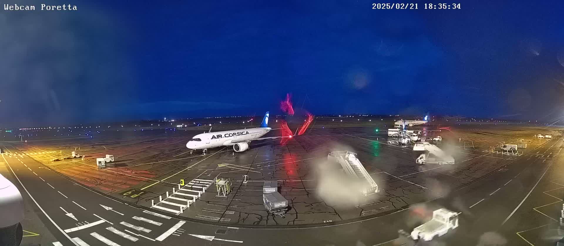 An airport tarmac at night under a dark blue, clear sky shows an Air Corsica plane parked near a jet bridge with airport vehicles and other planes in the distance.