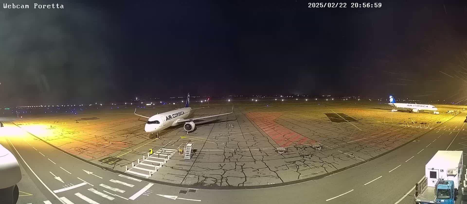 At night, a tarmac shows two airplanes parked and service vehicles, with runway lights visible in the distance under a dark sky.