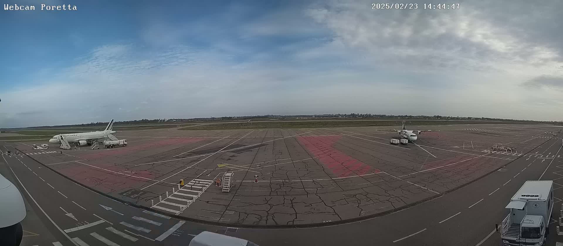 An airport tarmac under a partly cloudy sky shows two airplanes parked, one larger than the other, with ground support vehicles nearby.