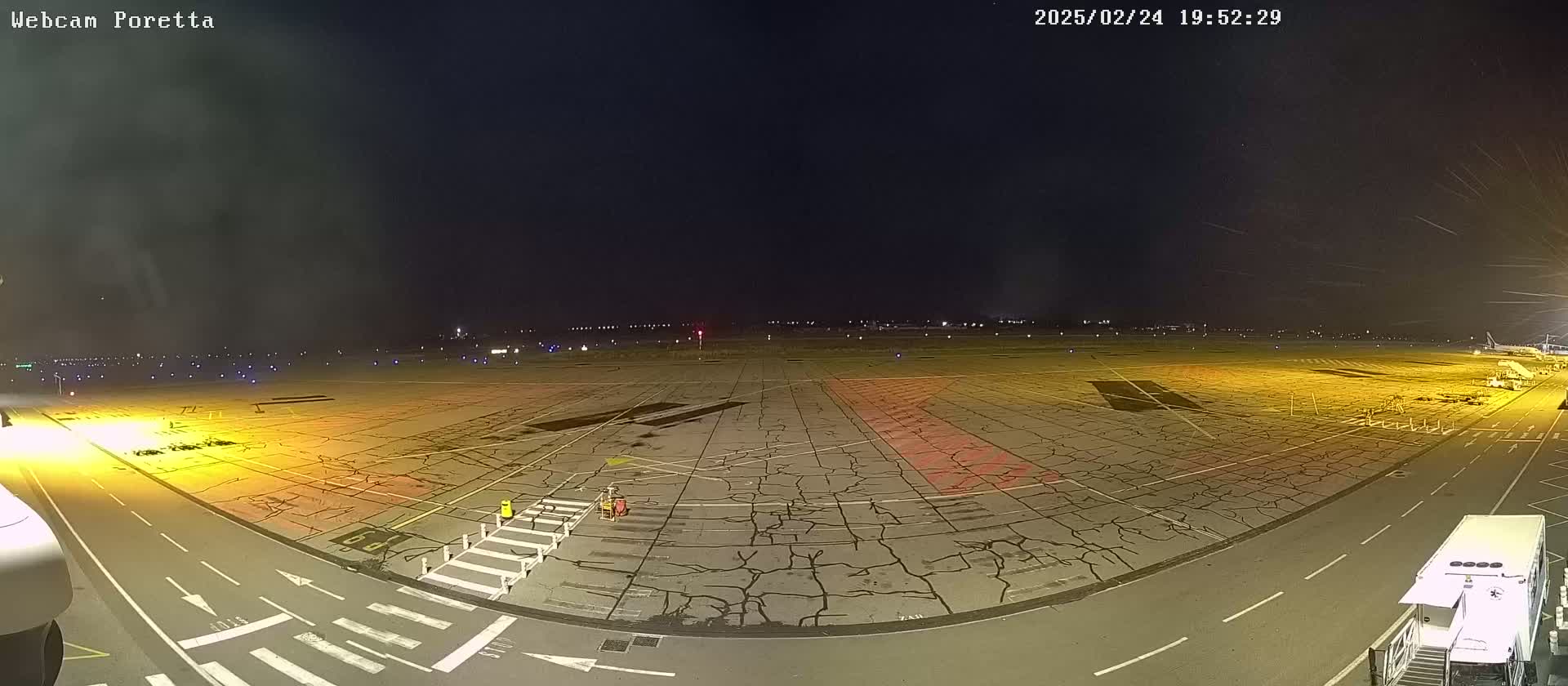 A nighttime view of an airport tarmac, showing a cracked surface with various markings and a few vehicles present under a dark sky.