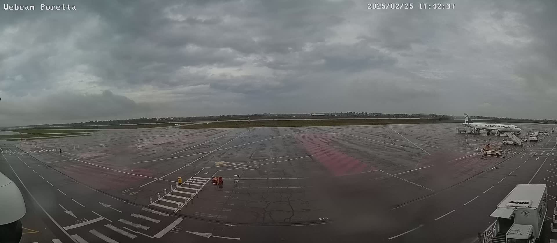 A wet airport tarmac under a cloudy sky shows an airplane parked at the gate with ground support equipment.