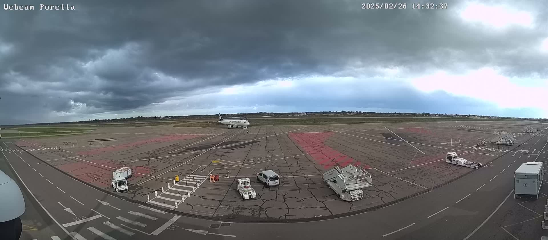 An airport tarmac under a cloudy sky shows an airplane and several ground service vehicles.
