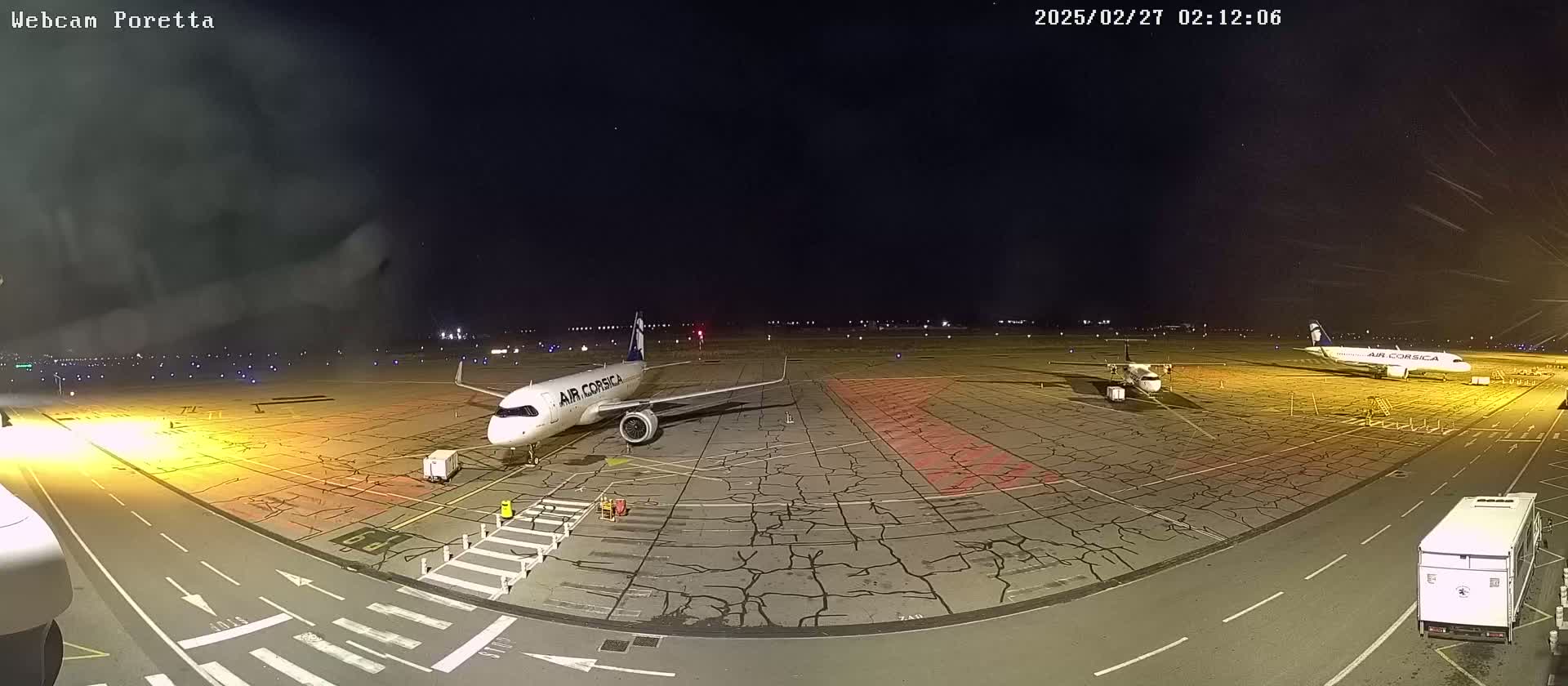 At night, an airport tarmac shows several airplanes parked, with one prominently displaying the Air Corsica logo; ground support vehicles are present.