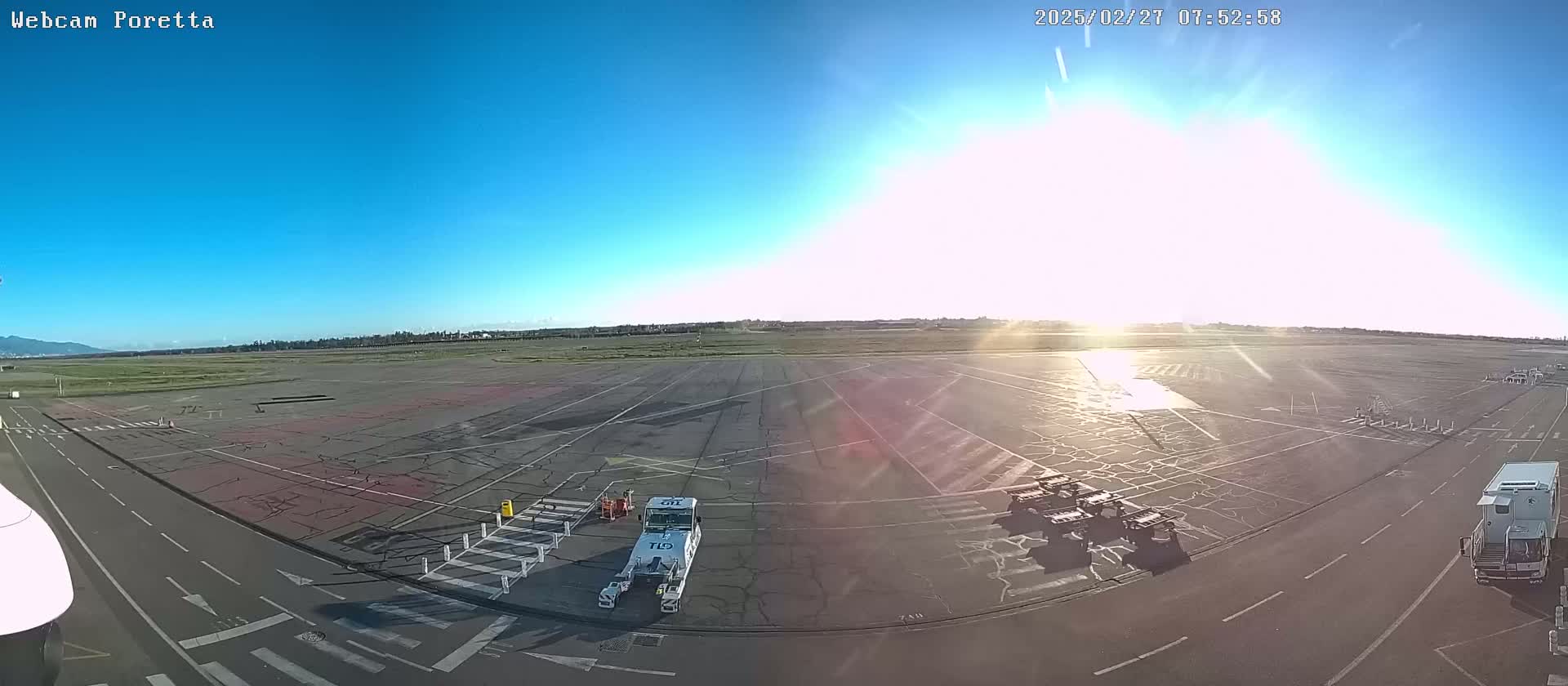 A sunny, clear day at an airport tarmac shows several ground support vehicles and a large expanse of cracked pavement.