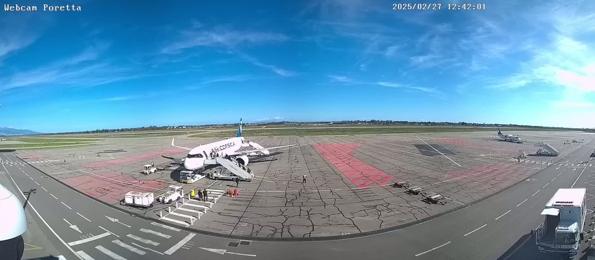 A passenger plane is parked at an airport tarmac under a mostly clear blue sky, with other planes and airport support vehicles visible.