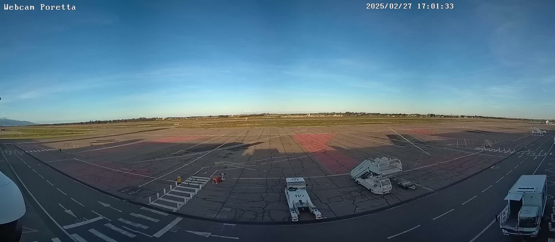 A mostly empty airport tarmac under a clear blue sky is seen from a high vantage point, with several ground service vehicles parked near the edge.