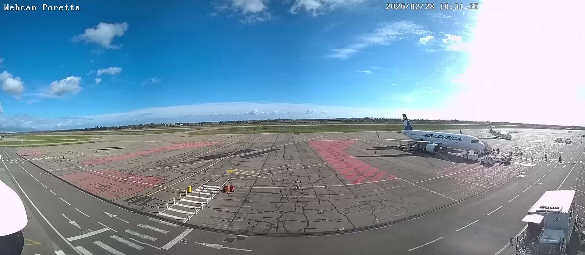 A mostly sunny day at an airport shows an Air Corsica airplane parked at a gate with passengers disembarking, other aircraft in the distance, and service vehicles on the tarmac.