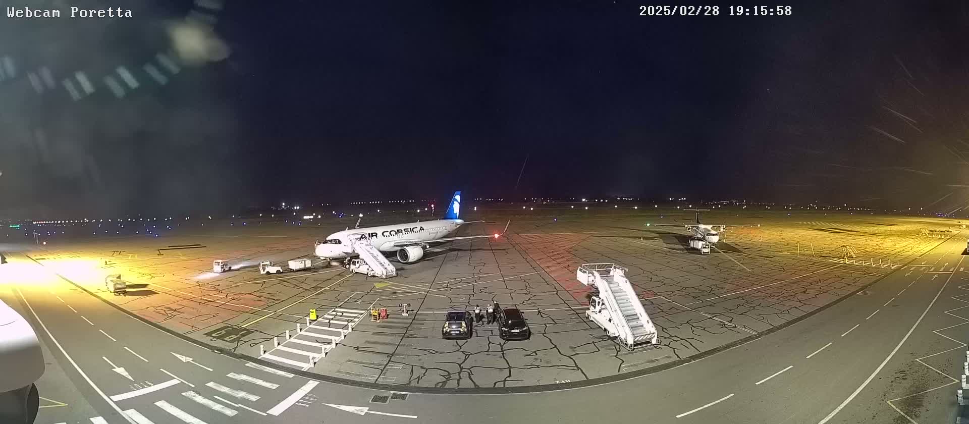 A nighttime view of an airport tarmac showing an Air Corsica airplane at a gate with a jet bridge attached, another smaller plane, and several ground service vehicles.