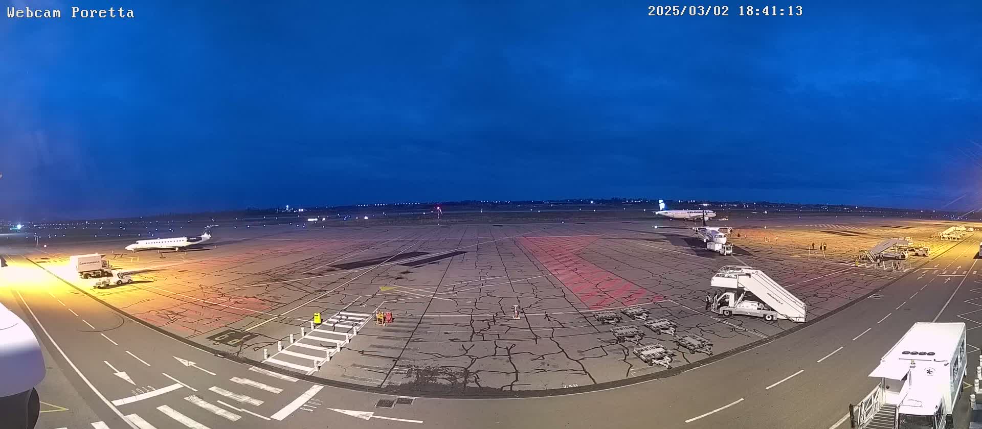 An airport tarmac at twilight, showing two airplanes parked with jet bridges attached, ground support equipment, and a mostly clear, dark blue sky.