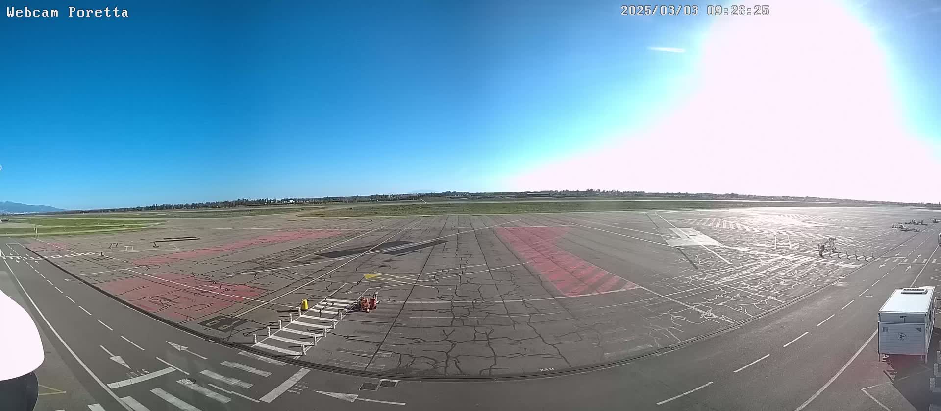 A sunny day at an airport tarmac shows a large expanse of cracked asphalt with markings, a few vehicles, and a clear blue sky.