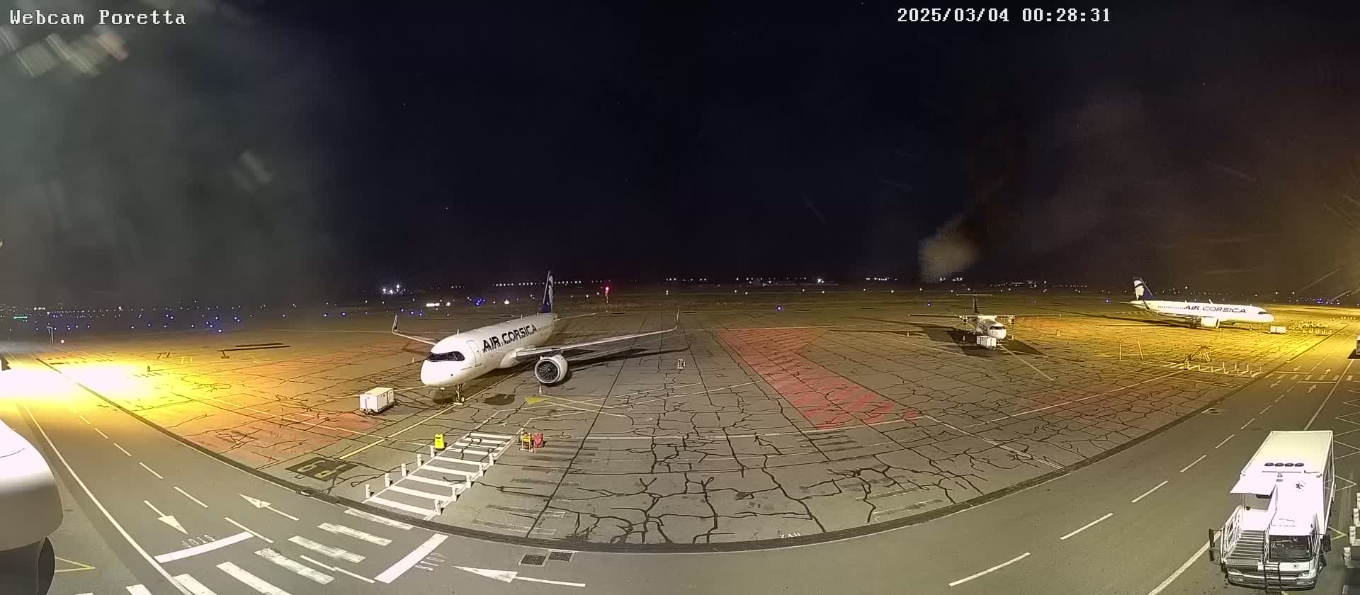 A nighttime airport tarmac shows two airplanes parked, one larger than the other, with various ground support vehicles present under a clear, dark sky.