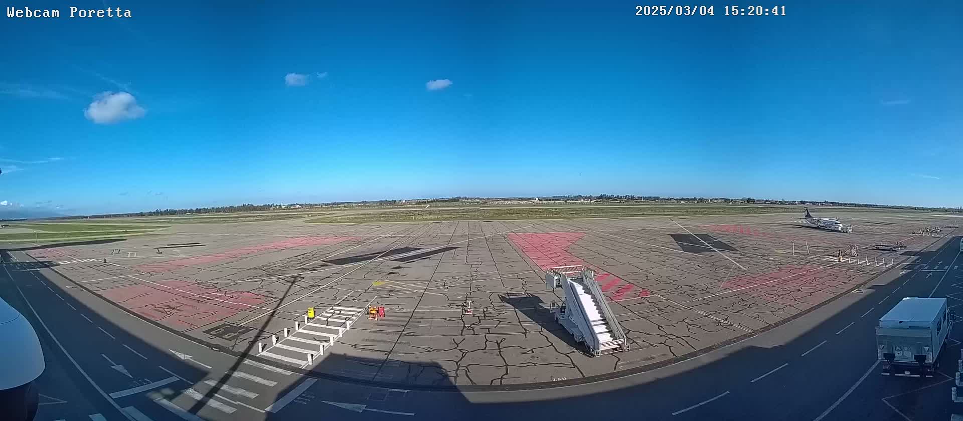 A mostly sunny airfield with a single airplane parked near a jet bridge and a few ground support vehicles.