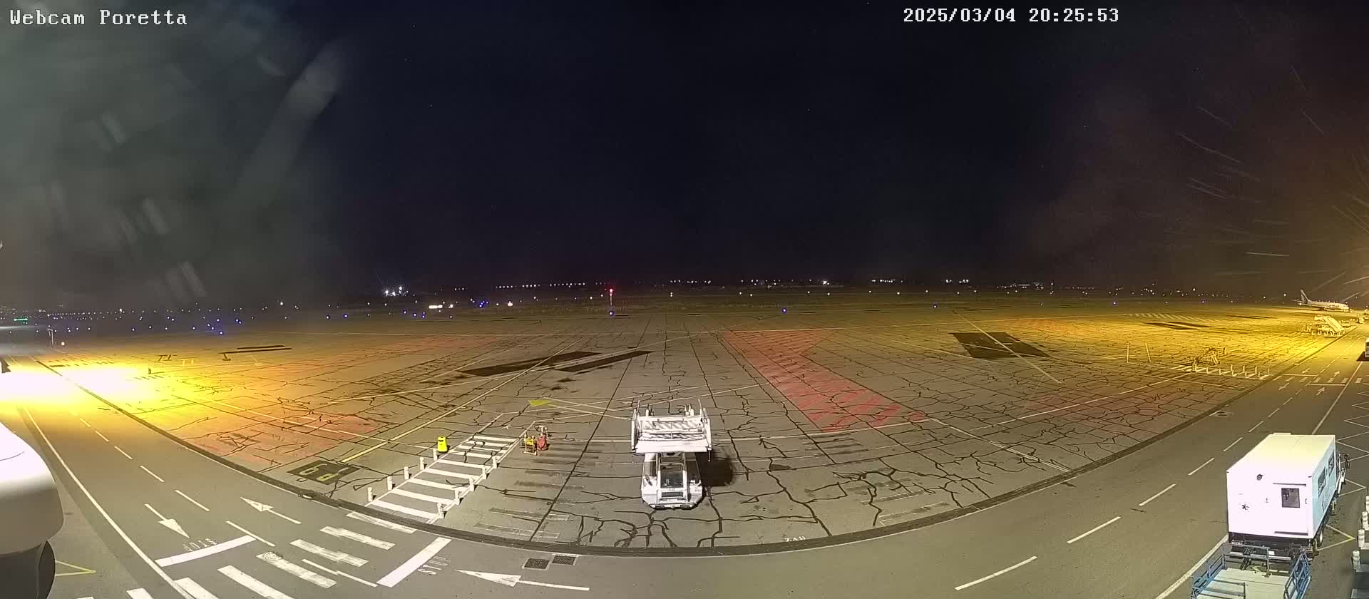 A nighttime view of an airport tarmac showing a jet bridge and ground support vehicles on a cracked asphalt surface under a clear, starry sky.
