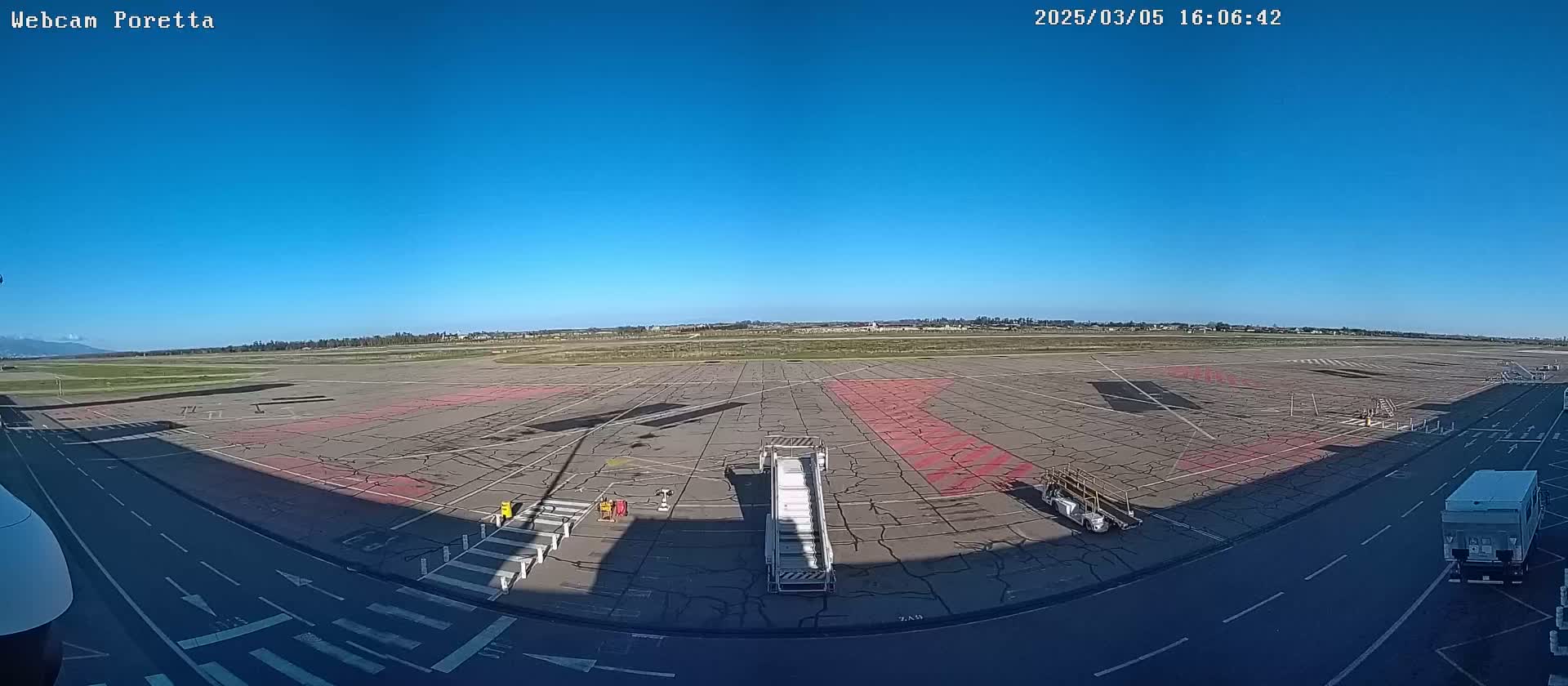A clear, sunny day at an airport tarmac shows jet bridges, ground support equipment, and a large expanse of cracked pavement.