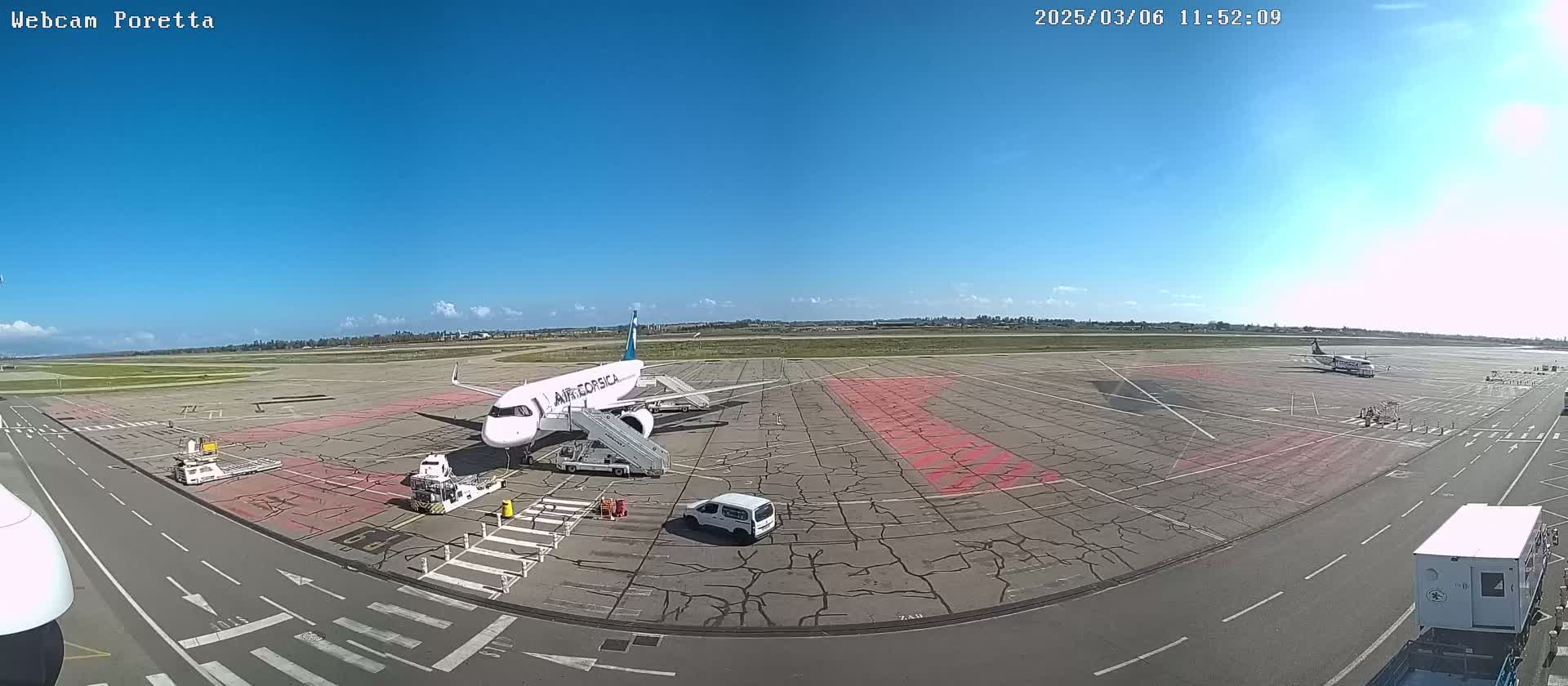A mostly sunny day at an airport shows an airplane at a gate with ground support vehicles, another smaller plane further away, and a few other vehicles.