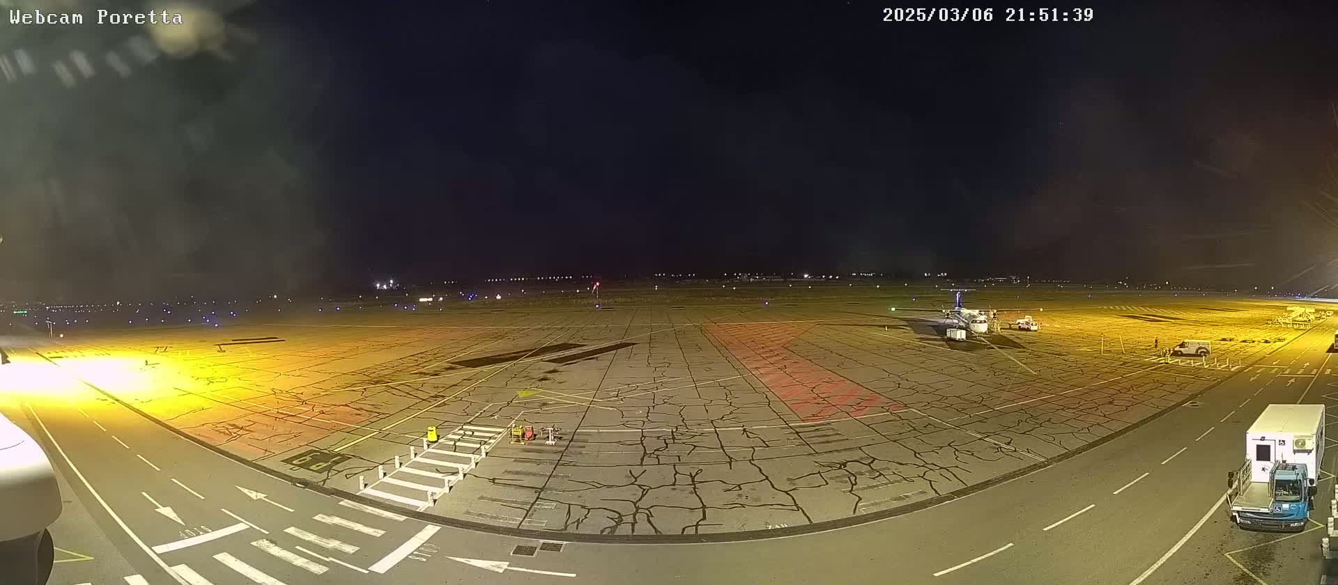 A nighttime view of a cracked tarmac airport apron, illuminated by artificial lights, with several vehicles and a small airplane parked on it.