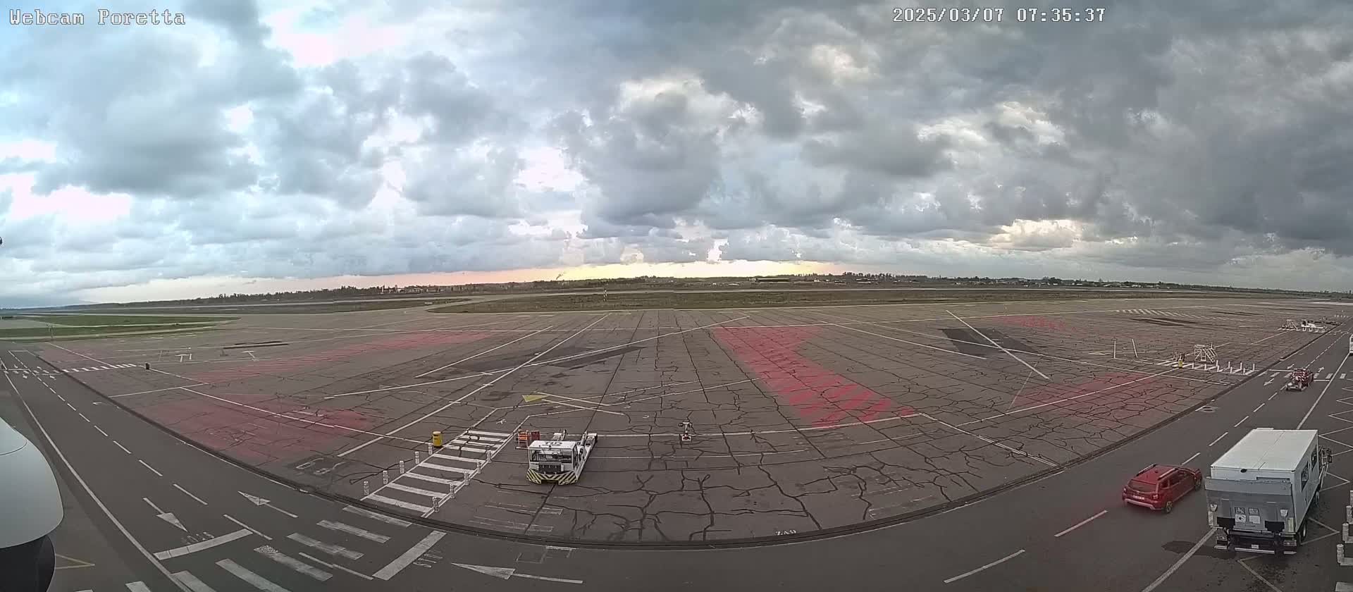 A large airport tarmac under a cloudy sky, with several vehicles and markings visible.