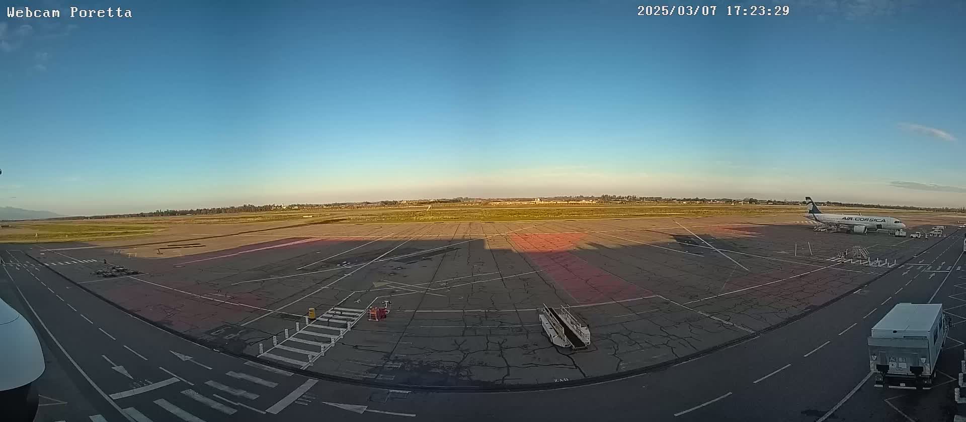 A wide shot of an airport tarmac under a clear blue sky shows an airplane parked near a baggage truck and other service vehicles.