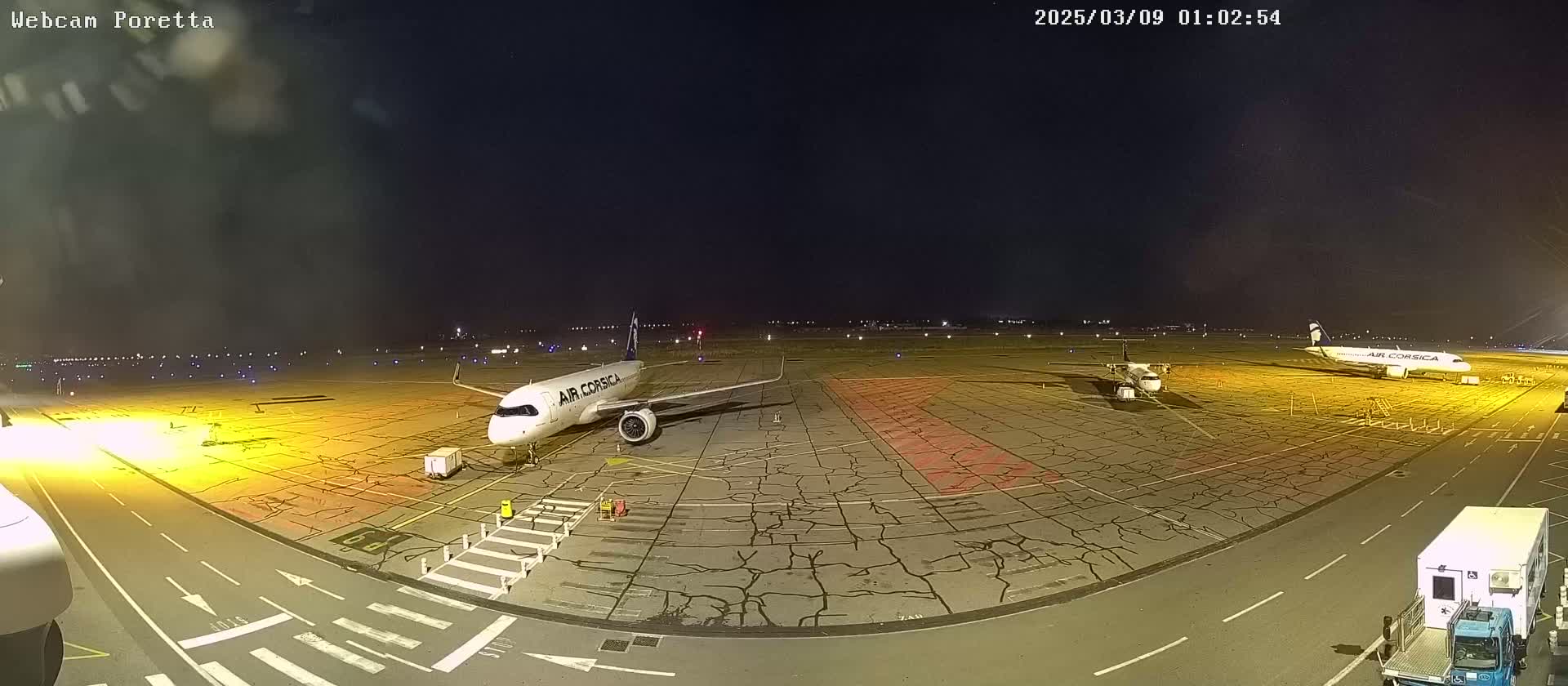 At night, two airplanes are parked on a tarmac with cracked pavement, surrounded by airport ground support vehicles.