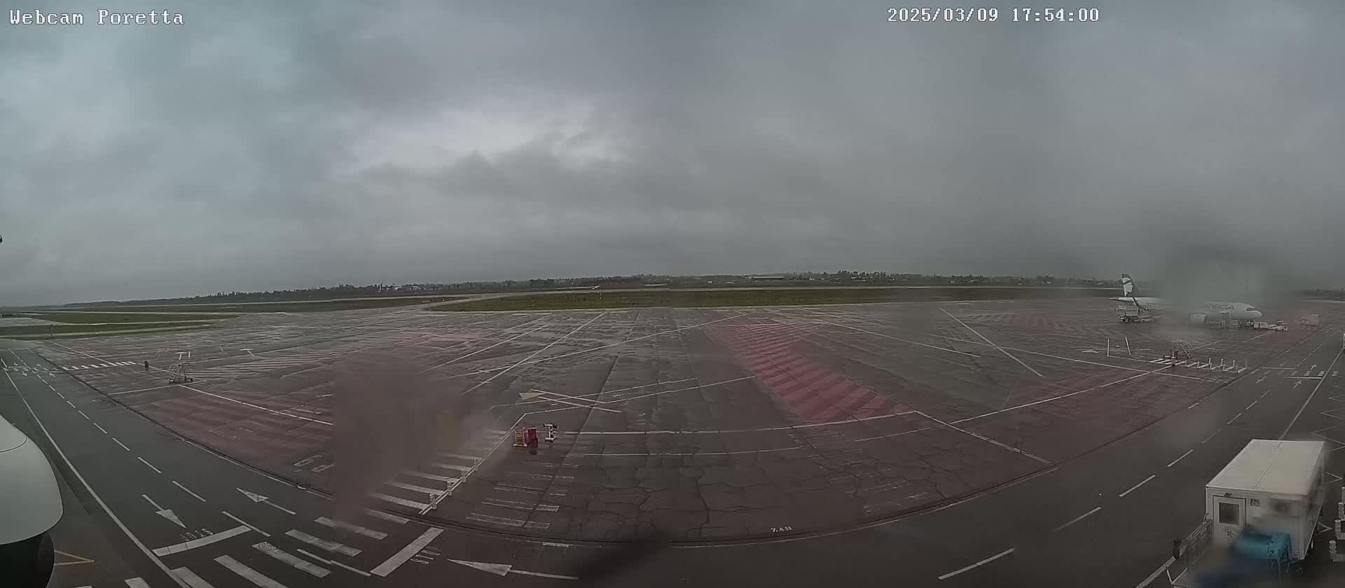 A wet airport tarmac under an overcast sky shows a parked airplane with ground support vehicles nearby.
