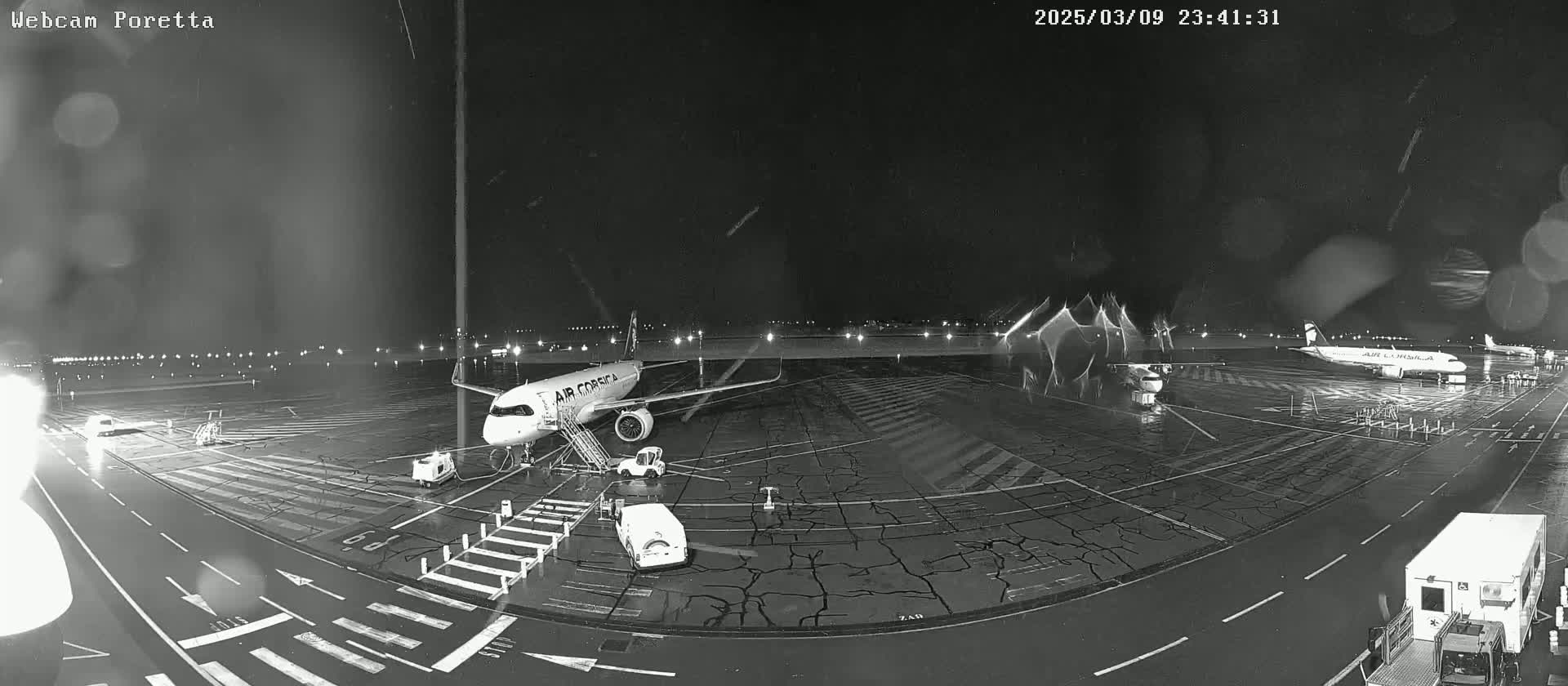 A nighttime airport tarmac shows an Air Corsica airplane at a gate with service vehicles present, under rainy conditions.