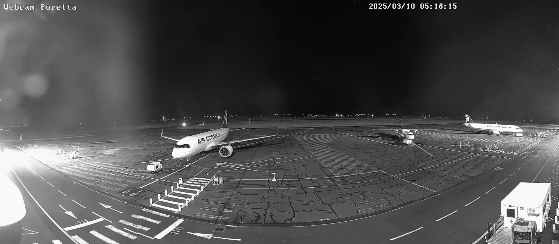 A nighttime, black-and-white view of an airport tarmac showing two airplanes parked, one larger than the other, with ground support vehicles nearby.