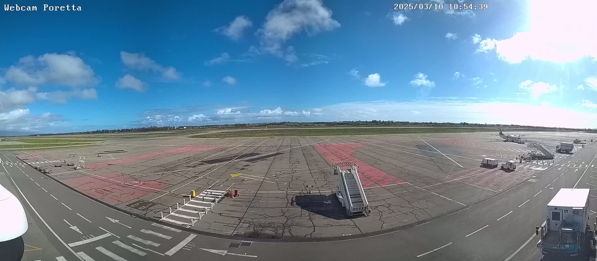 A mostly sunny day at an airport tarmac shows several service vehicles, a passenger jet, and a jet bridge.