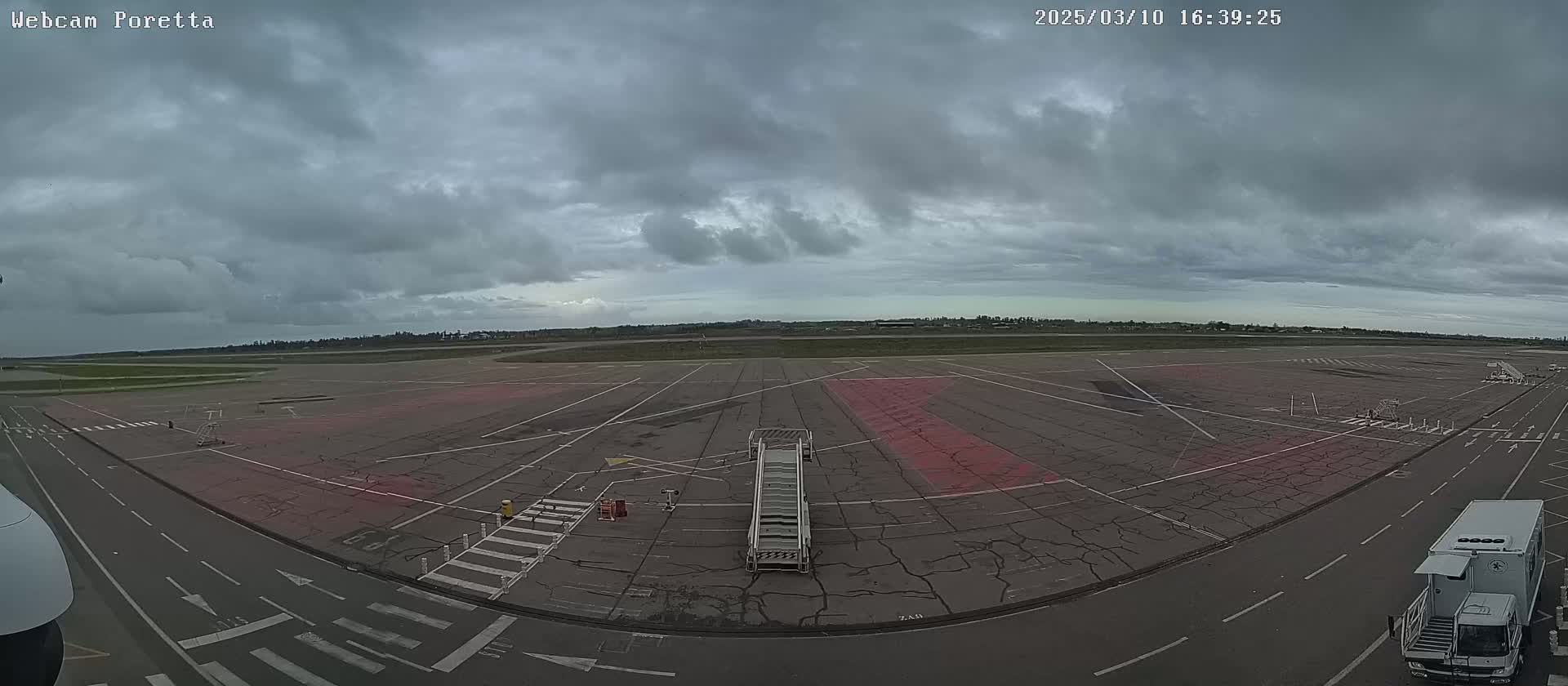 An overcast sky hangs over an empty airport tarmac with a jet bridge and airport service vehicles visible.