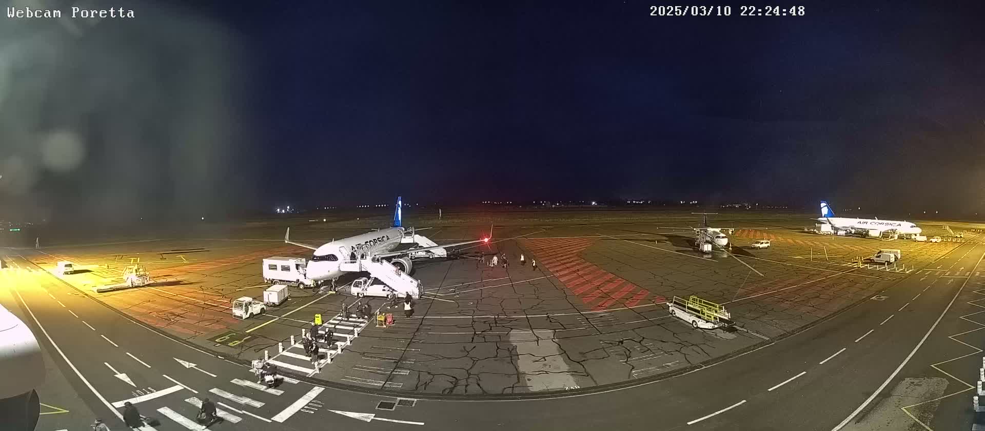 At night, under a clear, dark sky, two airplanes are parked at an airport tarmac with ground crews and vehicles present.