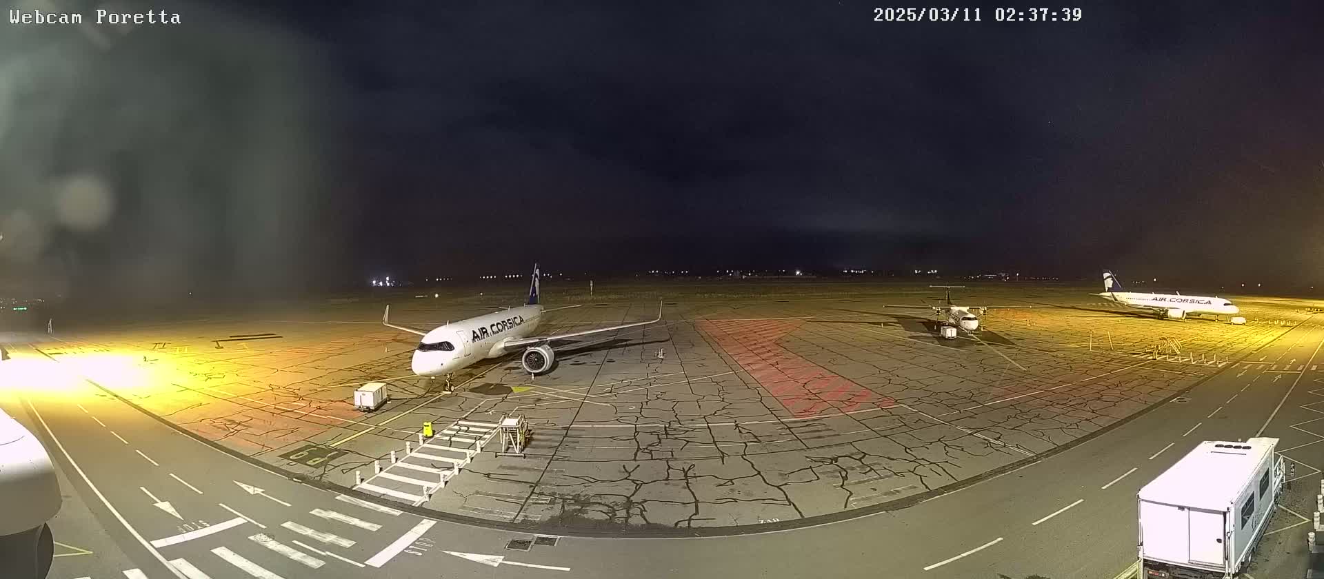 A nighttime view of an airport tarmac shows two airplanes parked, one larger than the other, with ground support vehicles nearby under a dark, cloudy sky.