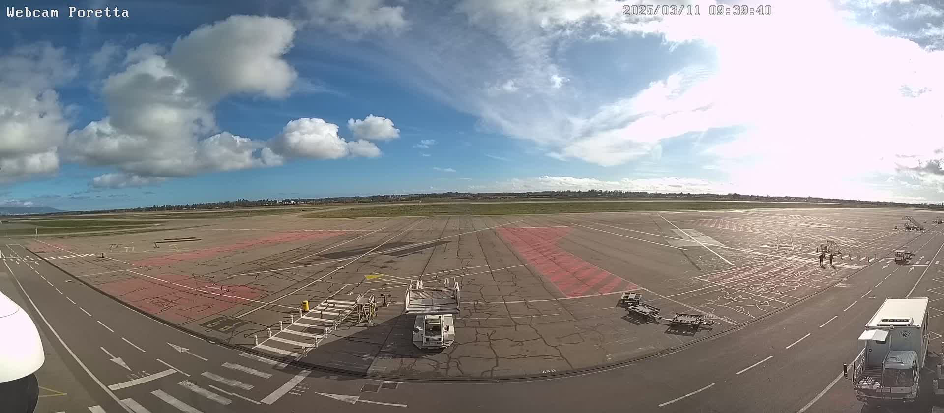 A mostly sunny day at an airport tarmac shows several ground service vehicles, with a few people visible in the distance and some cumulus clouds in the sky.