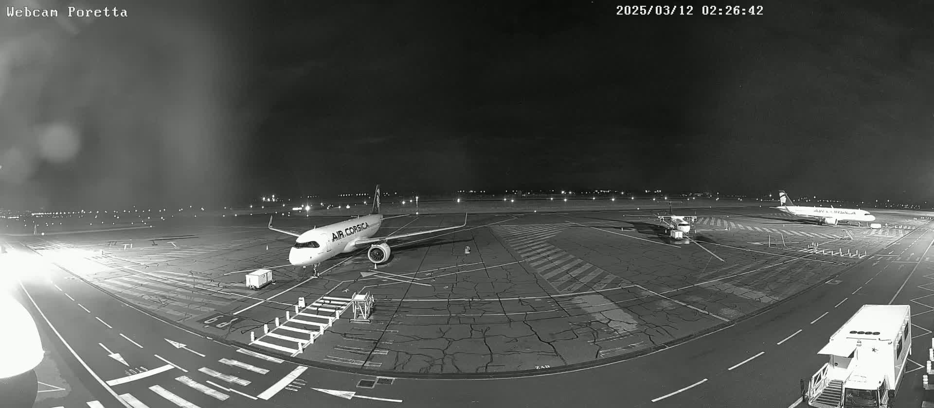 A nighttime view of an airport tarmac shows two airplanes parked, one of which is an Air Corsica plane, with airport ground support vehicles present under a dark sky.