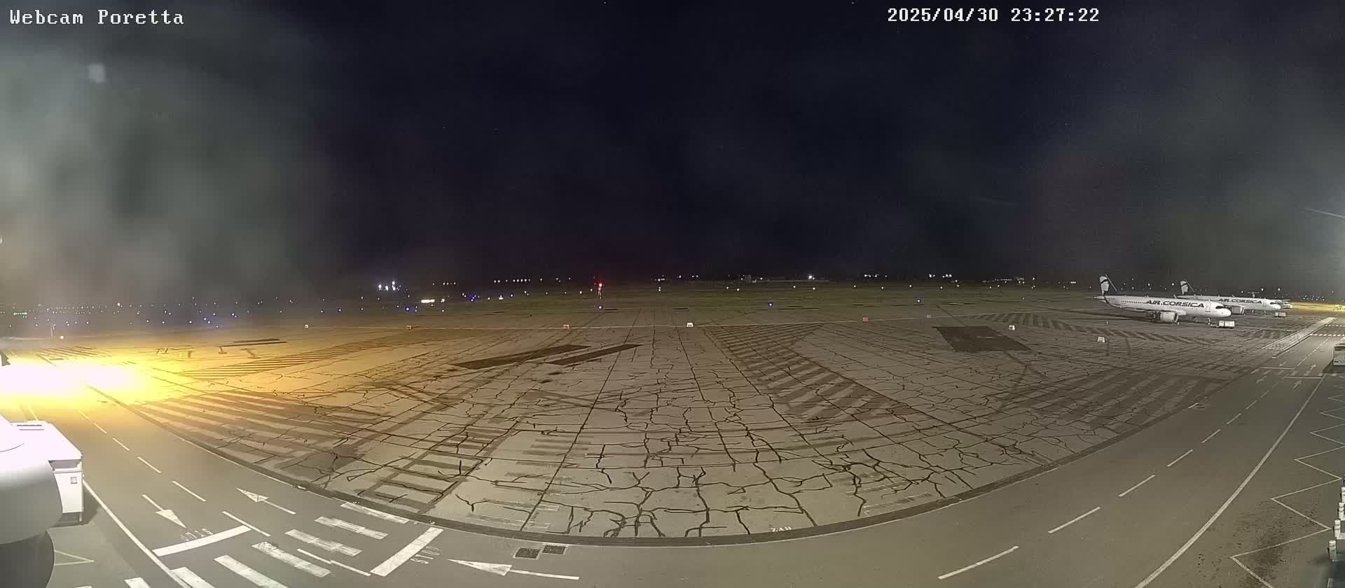 A nighttime view of an airport tarmac shows two airplanes parked near the edge, with a cracked asphalt surface extending to a runway lit with lights in the distance under a dark, clear sky.