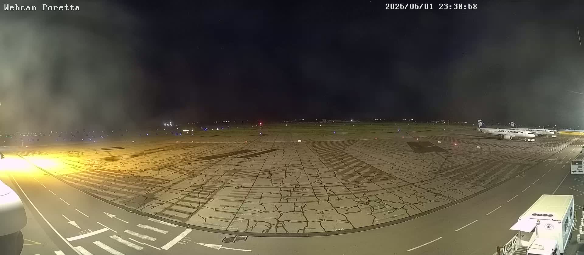 A nighttime view of an airport tarmac shows two airplanes parked near the edge, with a cracked surface and faint fog or haze in the distance.
