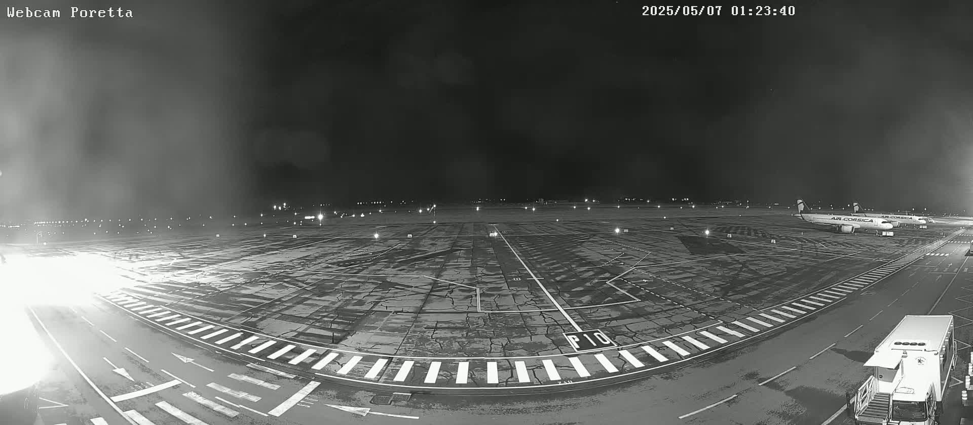 A nighttime, black and white view of an airport tarmac with two airplanes parked, a few service vehicles, and wet pavement under a dark sky.