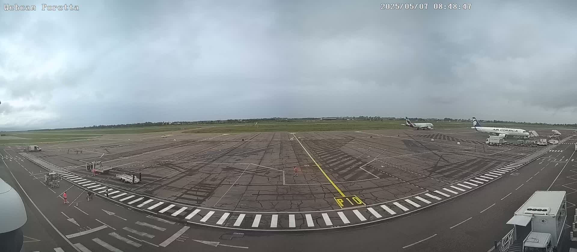 An overcast day at an airport shows tarmac, several planes, ground support vehicles, and a few people.