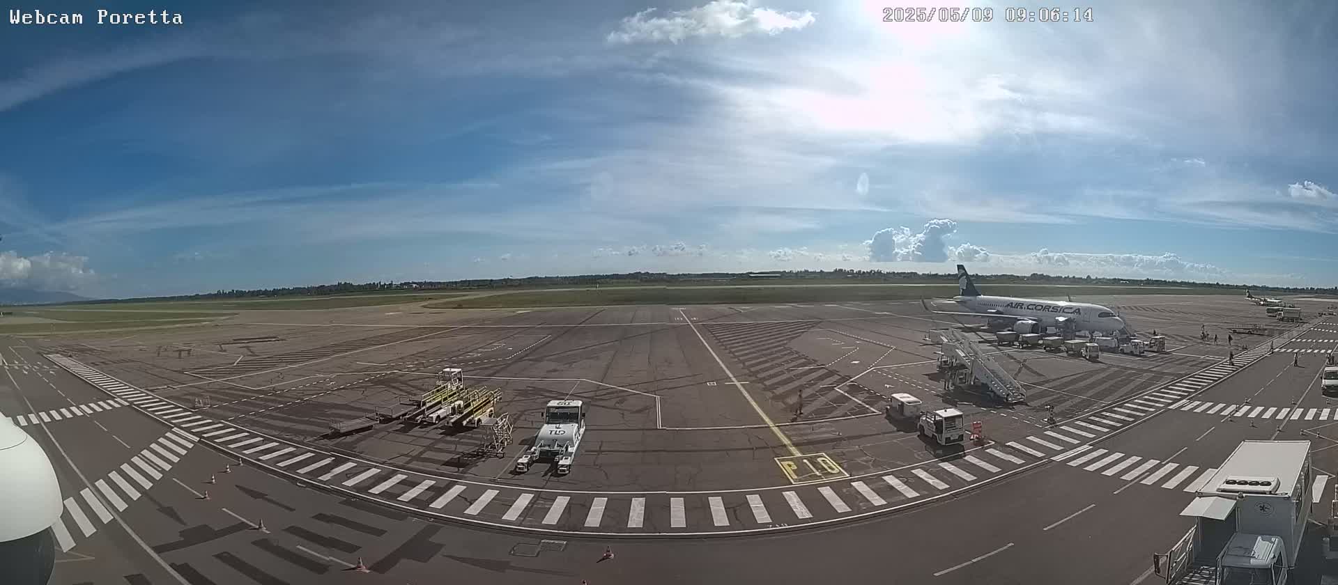 A mostly sunny day at an airport shows a passenger plane at a gate with ground support vehicles and other aircraft in the distance.
