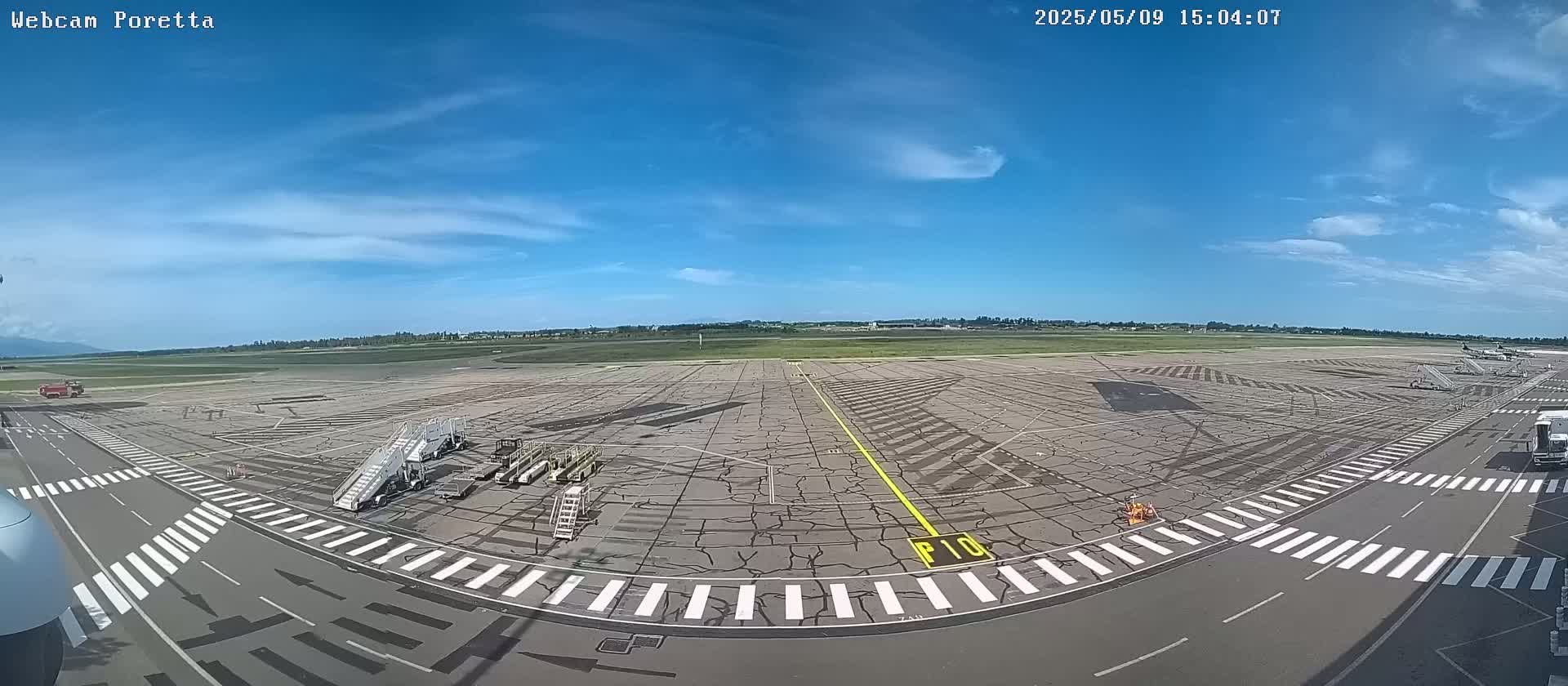 A wide, panoramic view of an airport tarmac on a sunny day with a few clouds, showing several parked airplanes and ground support equipment.