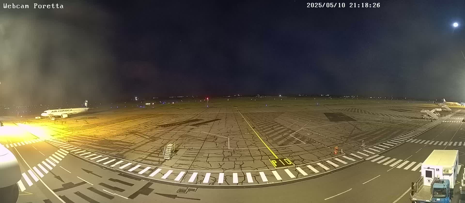 A nighttime view of an airport tarmac, showing an airplane parked at a gate, other ground support vehicles, and a mostly clear sky.