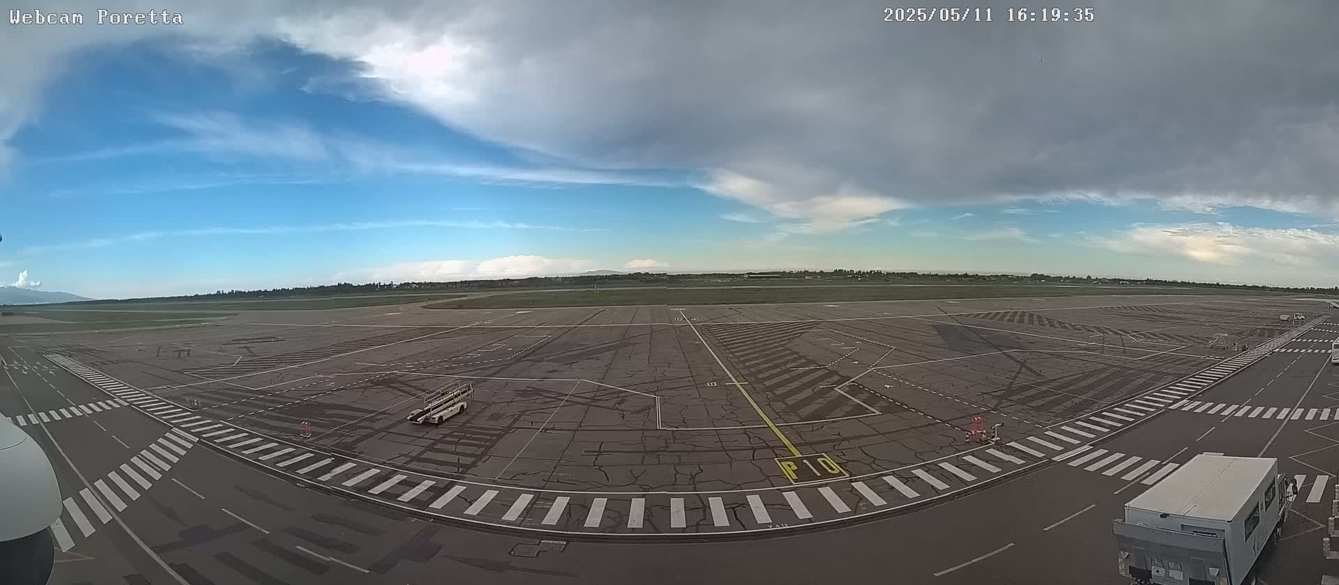 A mostly sunny airport tarmac with some clouds in the sky shows several vehicles and markings on the ground.