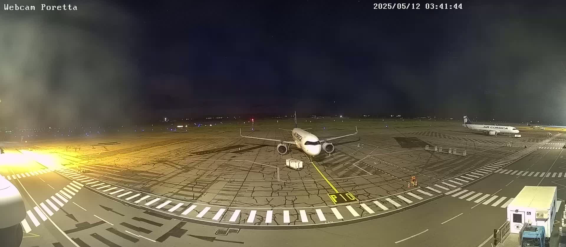 A nighttime view of an airport tarmac shows two airplanes parked, one closer to the camera, with a slightly hazy atmosphere.