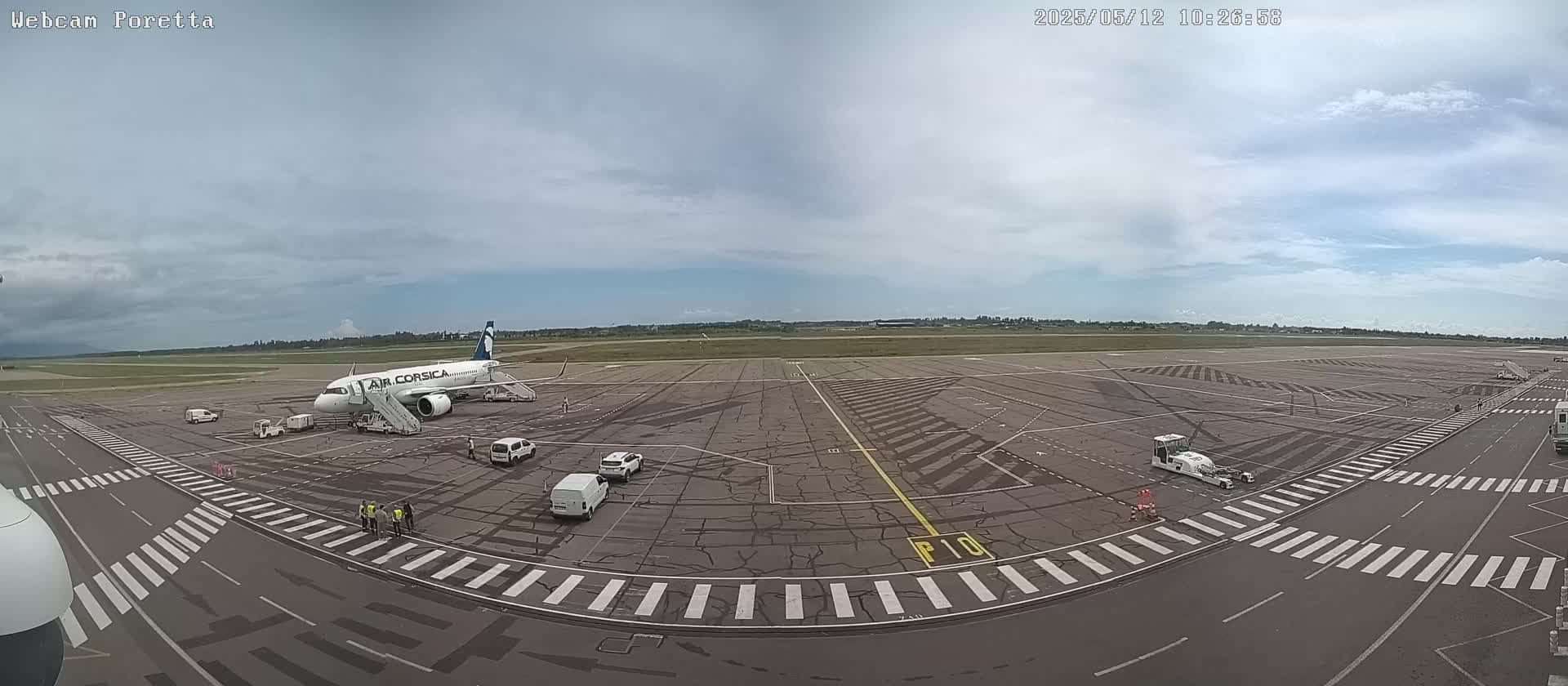 A passenger airplane is parked at an airport tarmac under a partly cloudy sky, with several ground vehicles and personnel nearby.