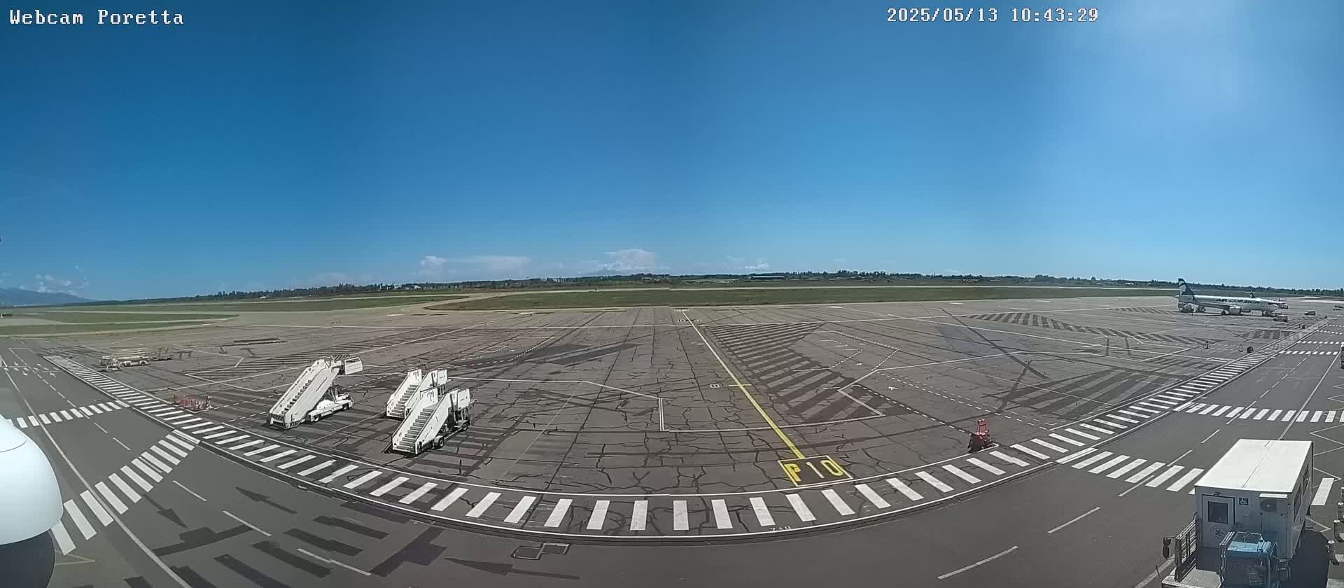 A wide, sunny outdoor shot shows an airport tarmac with several airplanes, jet bridges, and ground support vehicles under a clear blue sky.