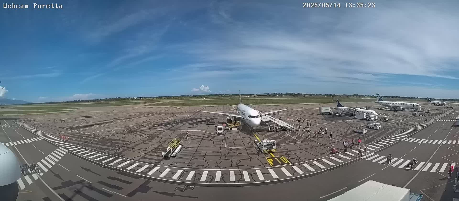 An airport tarmac on a partly sunny day shows several airplanes parked at gates with passengers and ground crews visible.